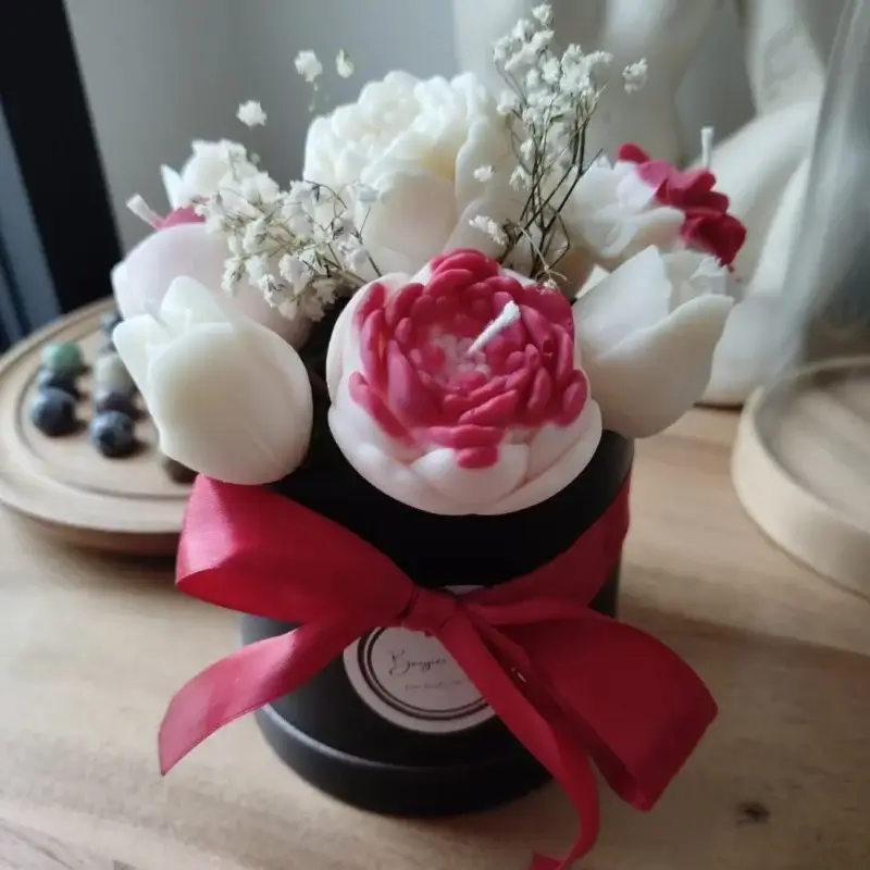 Bouquet de bougies artisanales dans les tons rouges et blanc disposées dans un pot rond en carton noir orné d'un ruban soyeux rouge.