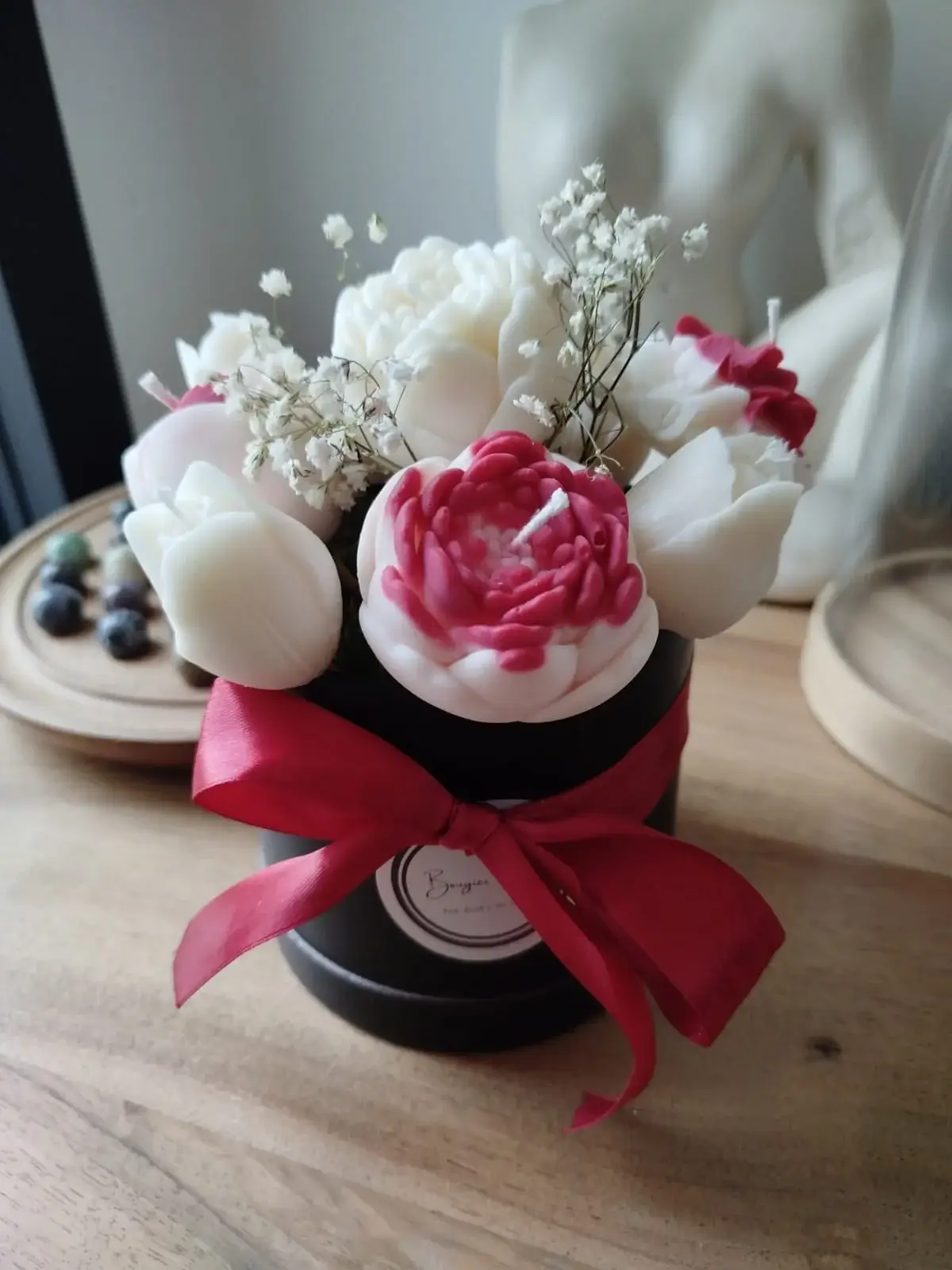Bouquet de bougies artisanales dans les tons rouges et blanc disposées dans un pot rond en carton noir orné d'un ruban soyeux rouge.